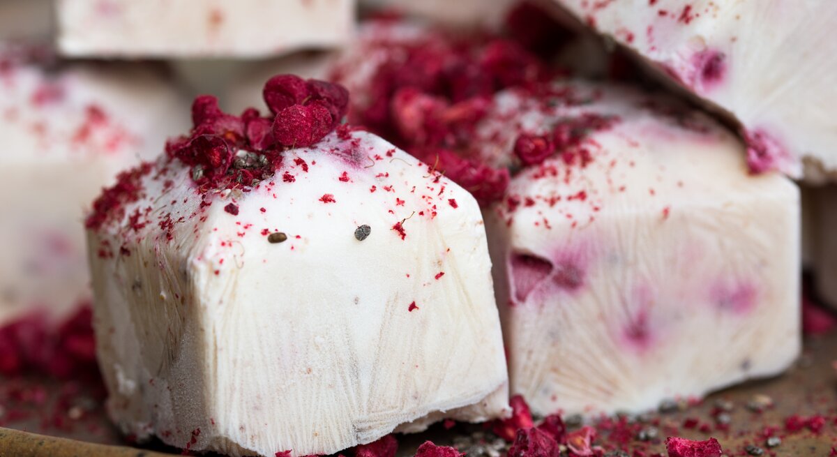 Mehrere Frozen Himbeer-Joghurt-Würfel liegen auf einem Untergrund auf Holz. Es handelt sich um gefrorene Sahne-Joghurt-Würfel mit Himbeeren.