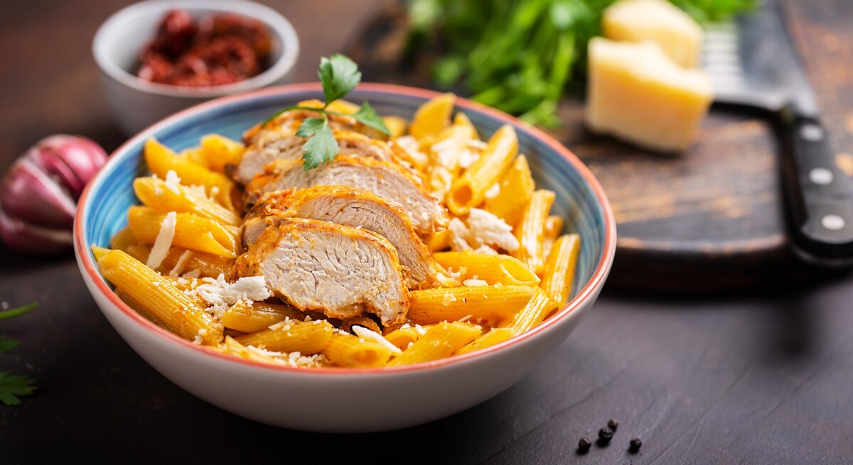 Cajun One-Pot-Pasta mit Hähnchen in einem bunten tiefen Teller. Dahinter eine Schale mit Tomaten, Gewürze und Parmesan. 