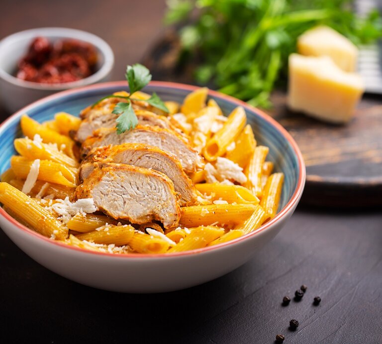 Cajun One-Pot-Pasta mit Hähnchen in einem bunten tiefen Teller. Dahinter eine Schale mit Tomaten, Gewürze und Parmesan.  | © Getty Images/ VadimZakirov