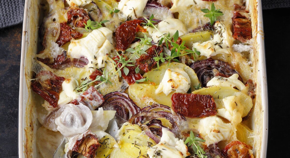Kartoffelgratin mit getrockneten Tomaten, roter Zwiebel und Thymian, mit Käse überbacken in einer großen Auflaufform auf einem dunklen Tisch. 