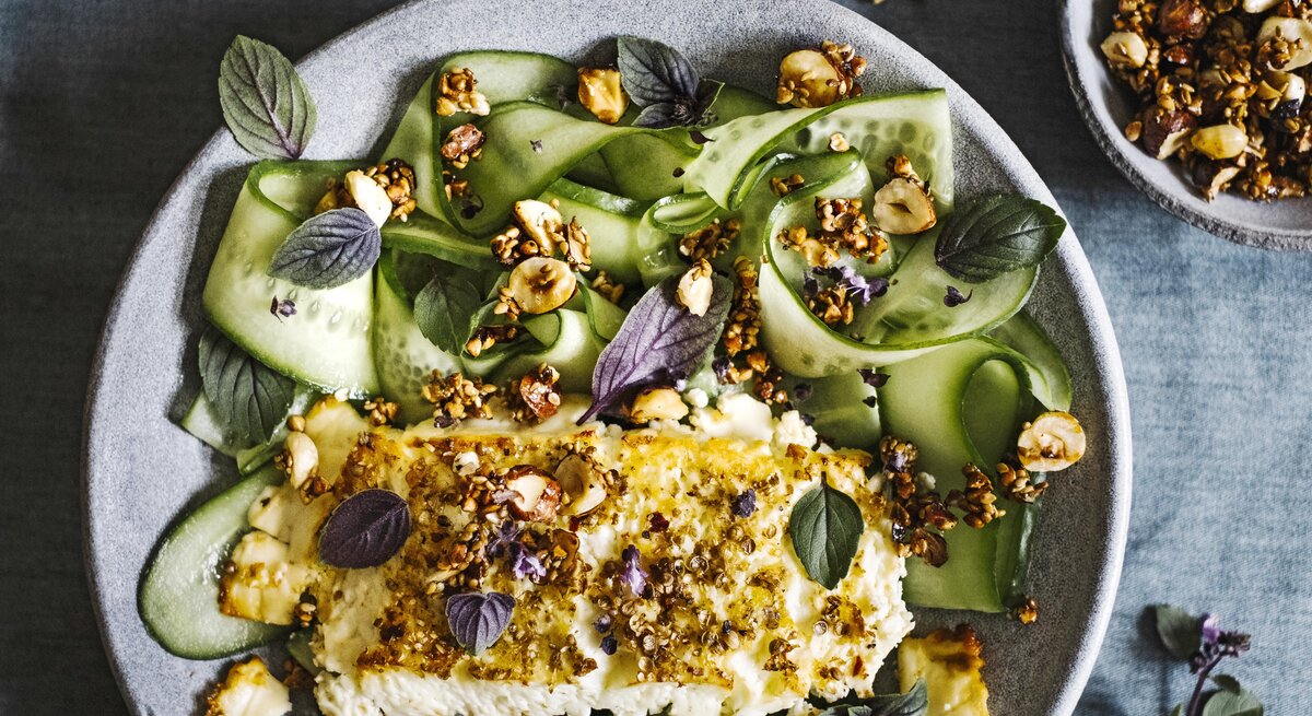 Gratinierter Feta mit Ahornsirup-Nusscrunch von oben mit Gurkengemüse in einem hellgrauen Teller neben einer Schüssel mit Nüssen auf einem grauen Tisch. 