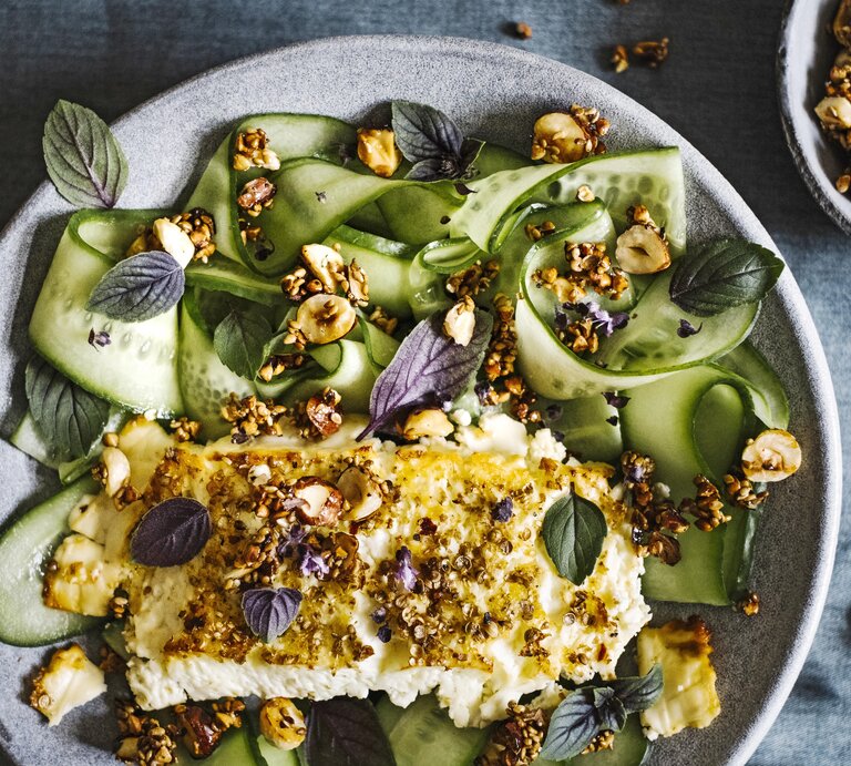 Gratinierter Feta mit Ahornsirup-Nusscrunch von oben mit Gurkengemüse in einem hellgrauen Teller neben einer Schüssel mit Nüssen auf einem grauen Tisch.  | © StockFood/Picture Press/Marina Jerkovic