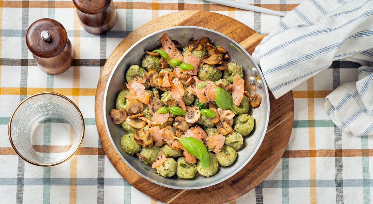 Draufsicht: Auf einem karierten Stoff stehen ein Salz- und ein Pfefferstreuer, ein Trinkglas, eine Gabel und ein rundes Holzbrettchen. Auf dem Brettchen steht eine Metallpfanne mit Bärlauch-Gnocchi mit Lachs.