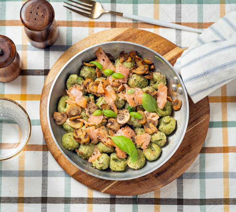 Draufsicht: Auf einem karierten Stoff stehen ein Salz- und ein Pfefferstreuer, ein Trinkglas, eine Gabel und ein rundes Holzbrettchen. Auf dem Brettchen steht eine Metallpfanne mit Bärlauch-Gnocchi mit Lachs. | © Getty Images / 	Carlo A
