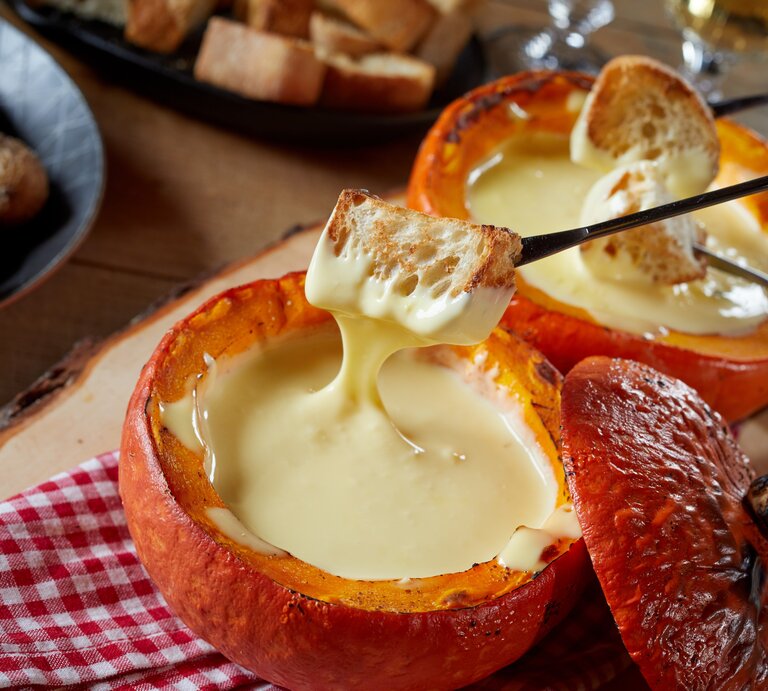 Käse-Fondue im Kürbis auf einem rustikalen Tisch mit rot-weiß-karierter Tischdecke. | © Shutterstock/ stockcreations