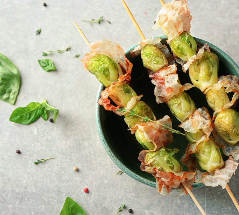 Drei Spieße mit Rosenkohl und Speck in einer Schüssel auf hellem Untergrund umgeben von Kräutern. | © Shutterstock/Africa Studio