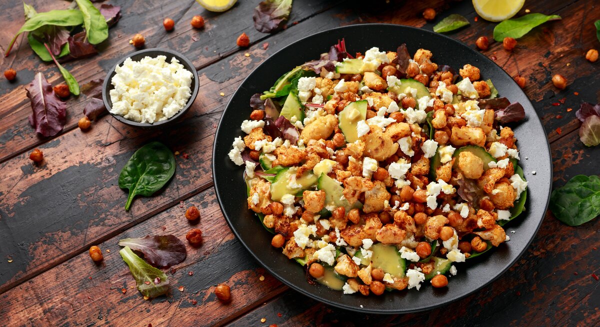 Eine große schwarze Schüssel mit dem gerösteten Blumenkohl-Kichererbsen-Salat mit Gurke und Feta neben einer kleinen Schüssel mit zerbröseltem Feta und vielen losen Salatblättern und Zitronenspalten, vor einem Holzhintergrund von schräg oben. 