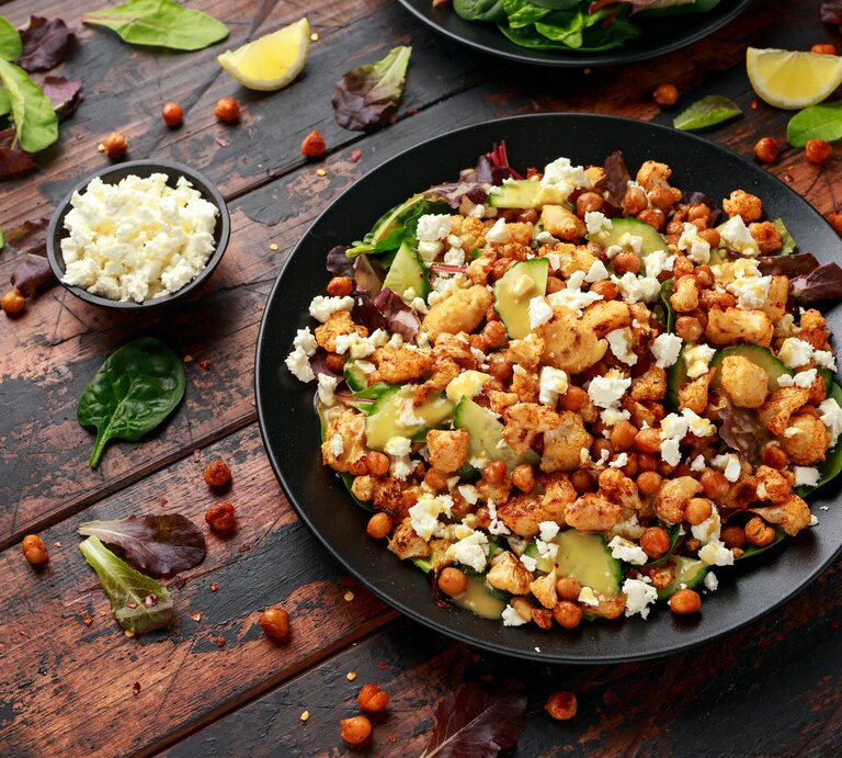 Eine große schwarze Schüssel mit dem gerösteten Blumenkohl-Kichererbsen-Salat mit Gurke und Feta neben einer kleinen Schüssel mit zerbröseltem Feta und vielen losen Salatblättern und Zitronenspalten, vor einem Holzhintergrund von schräg oben.  | © Getty Images/DronG