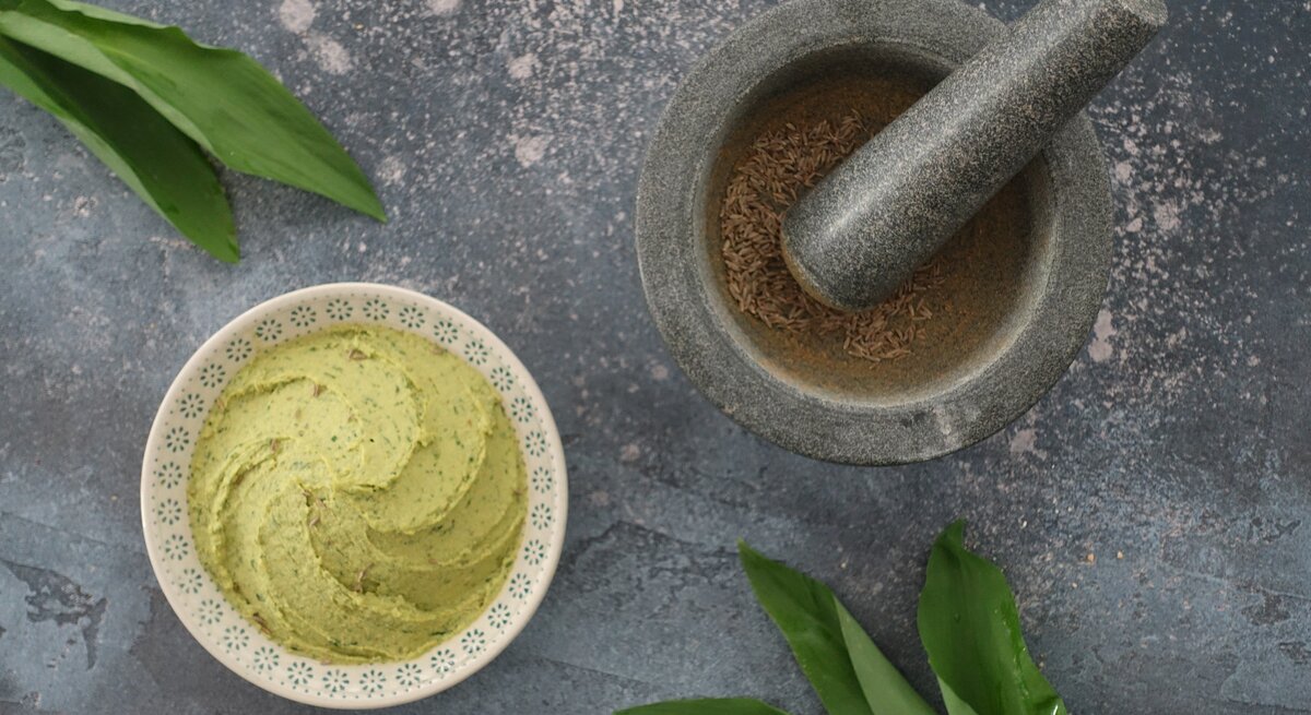Eine Schale Bärlauch-Hummus in der Draufsicht, daneben ein Mörser mit Pfeffer und einige Bärlauch-Blätter.