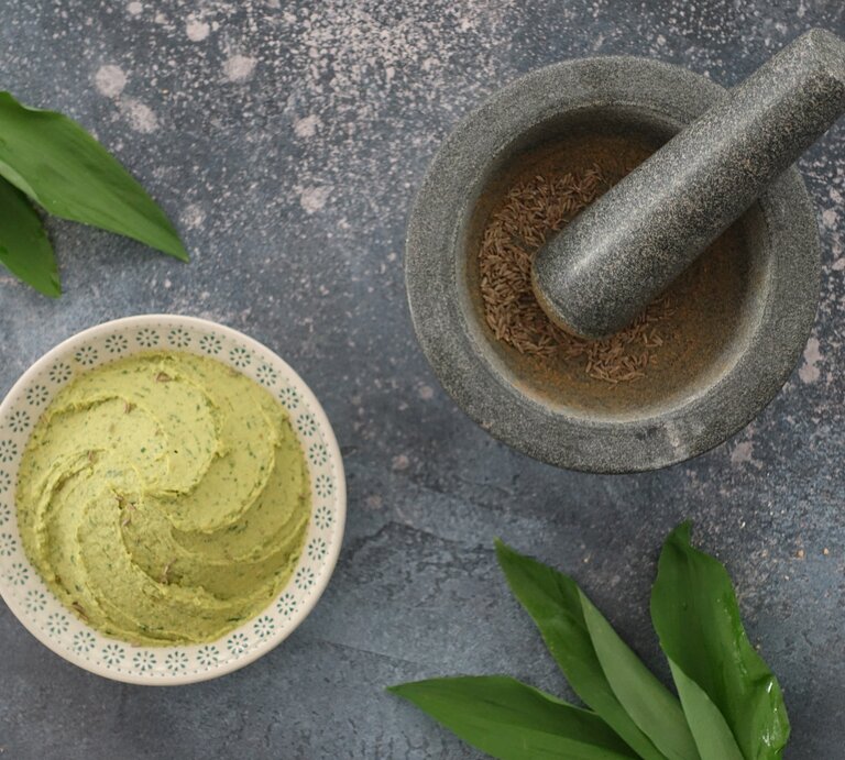 Eine Schale Bärlauch-Hummus in der Draufsicht, daneben ein Mörser mit Pfeffer und einige Bärlauch-Blätter. | © Shutterstock/ Paradieschen