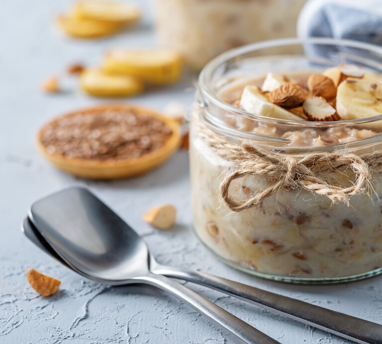 Ein Glas mit dem Leinsamen-Pudding auf hellblauem Untergrund. Daneben noch unscharf gehackte Nüsse und Bananenscheiben. | © Shutterstock/ Nataliya Arzamasova