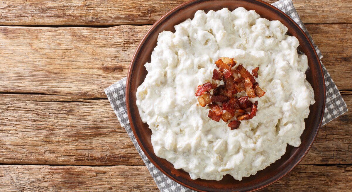 Auf einem Untergrund aus Holz liegt ein grau-weißes kariertes Tuch. Auf dem Tuch steht eine Schale mit Spätzle mit Feta und Speck. Es handelt sich um ein Gericht aud der Slowakei.