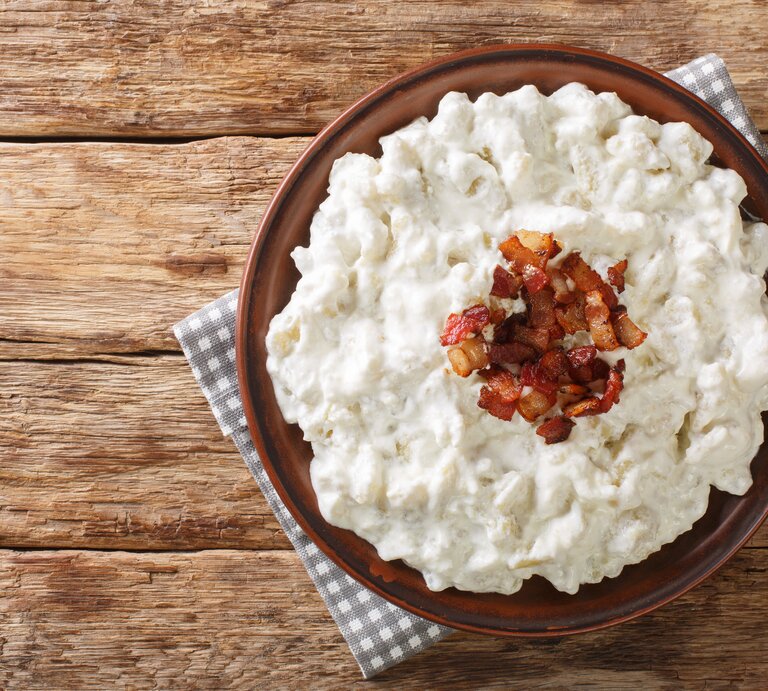 Auf einem Untergrund aus Holz liegt ein grau-weißes kariertes Tuch. Auf dem Tuch steht eine Schale mit Spätzle mit Feta und Speck. Es handelt sich um ein Gericht aud der Slowakei. | © Getty Images / ALLEKO