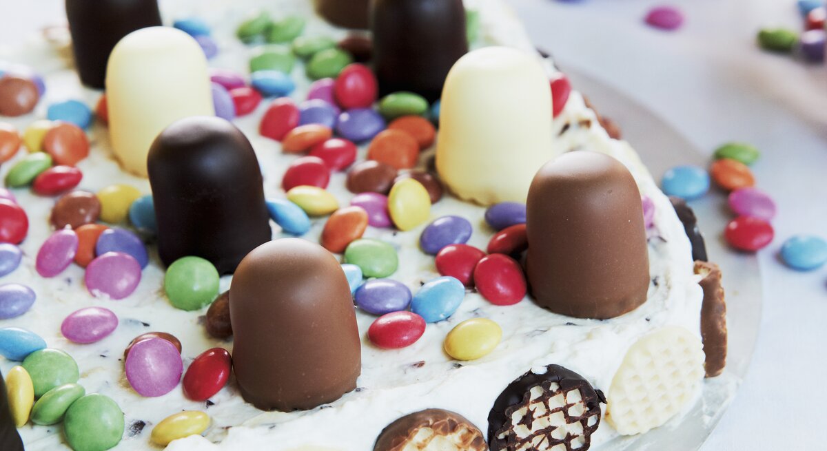 Auf einem Teller liegt eine bunte Mini-Schokokuss-Torte. Sie ist dekoriert mit Schokoküssen und Smarties. Im Hintergrund sind zwei Tassen zu sehen. 