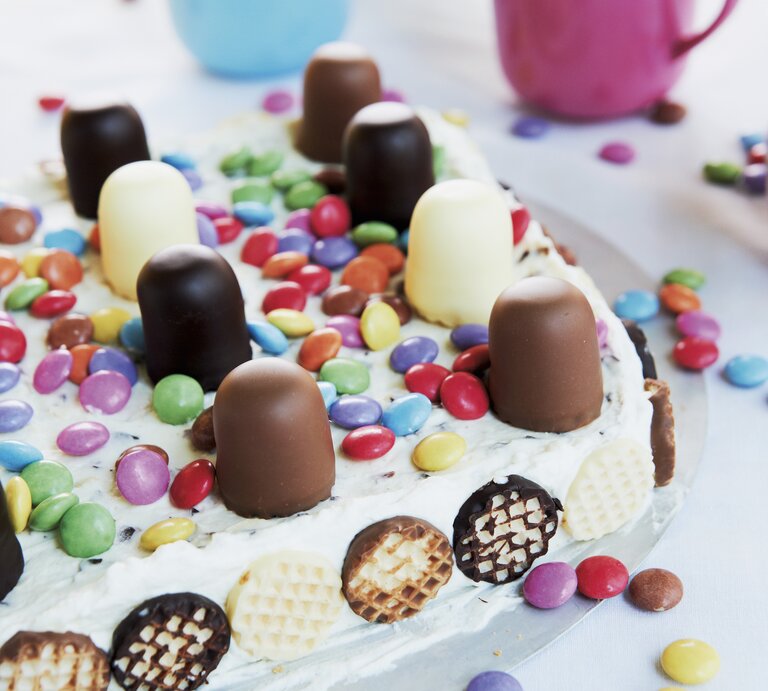 Auf einem Teller liegt eine bunte Mini-Schokokuss-Torte. Sie ist dekoriert mit Schokoküssen und Smarties. Im Hintergrund sind zwei Tassen zu sehen.  | © EAT CLUB/ Isabell Triemer