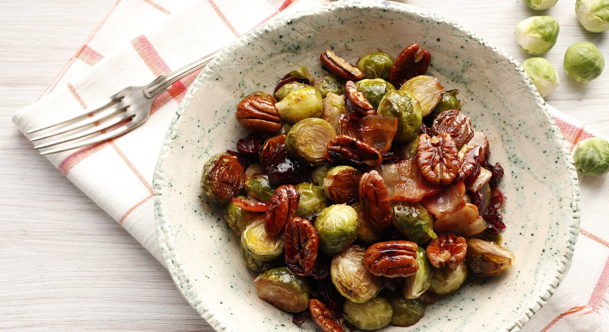 Ein Rosenkohl-Salat mit Nüssen und getrockneten Cranberries in einer weißen, gemusterten Schale steht auf einem karierten Geschirrtuch auf einem weißen Tisch. Neben der Schale liegt eine Gabel und einige einzelne Rosenkohlröschen.