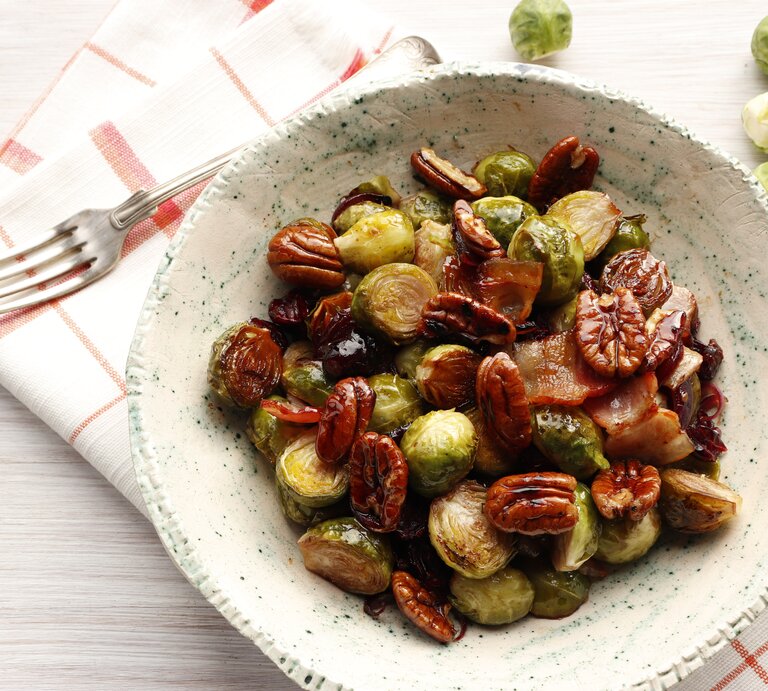 Ein Rosenkohl-Salat mit Nüssen und getrockneten Cranberries in einer weißen, gemusterten Schale steht auf einem karierten Geschirrtuch auf einem weißen Tisch. Neben der Schale liegt eine Gabel und einige einzelne Rosenkohlröschen. | © Shutterstock / Olga Bondarenko