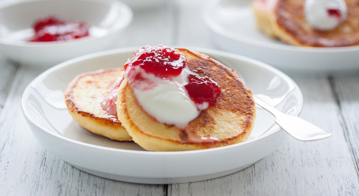 Hüttenkäse Pfannkuchen auf weißem Teller mit Joghurt und Himbeere verfeinert. Drei Portionen auf einem hellem Holztisch.