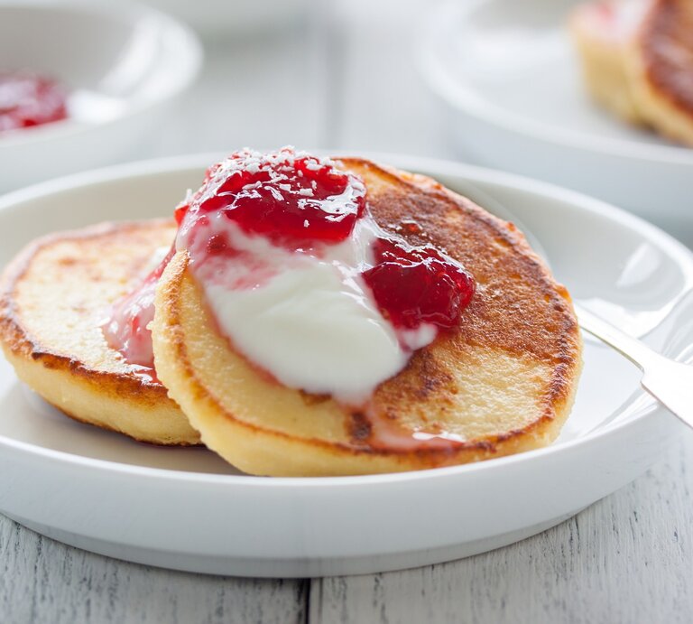 Hüttenkäse Pfannkuchen auf weißem Teller mit Joghurt und Himbeere verfeinert. Drei Portionen auf einem hellem Holztisch. | © Getty Images/iStockphoto
