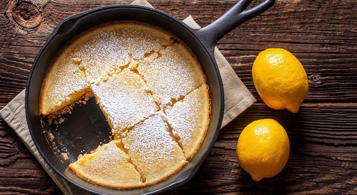 Zitronenkuchen ohne Backen aus der Pfanne in schwarzer Gusspfanne auf beigem Tuch und dunklem Holz. Daneben zwei Zitronen. Aufnahme aus der Vogelperspektive.