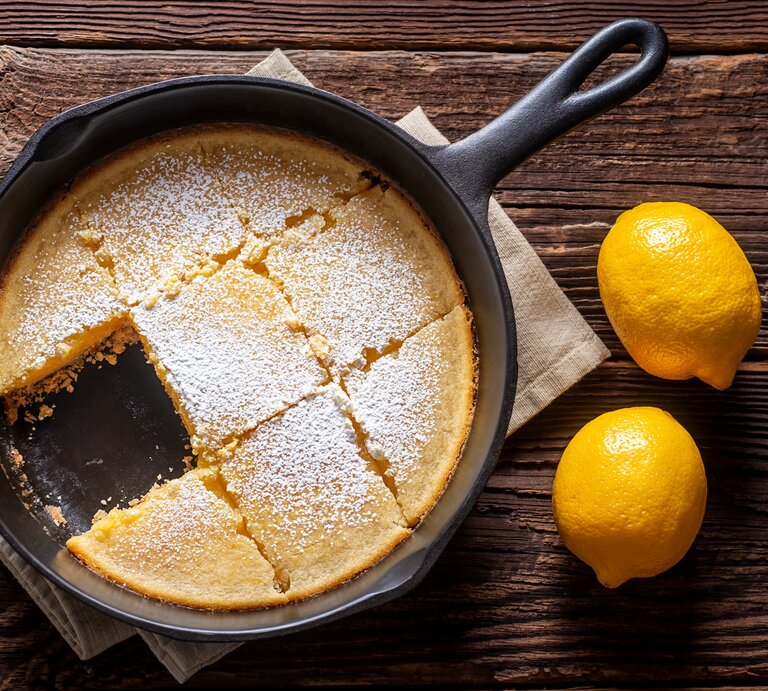 Zitronenkuchen ohne Backen aus der Pfanne in schwarzer Gusspfanne auf beigem Tuch und dunklem Holz. Daneben zwei Zitronen. Aufnahme aus der Vogelperspektive. | © Getty Images / rudisill