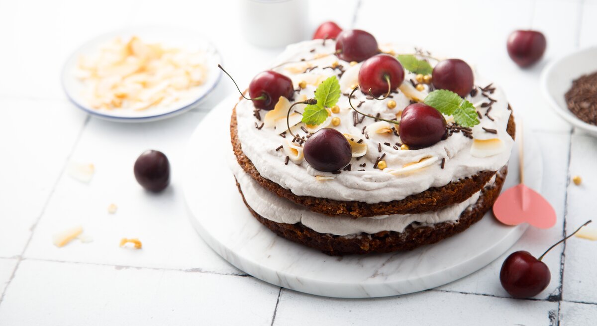 Schüttelkuchen nach Schwarzwälder-Kirsch-Art auf weißem, hölzernem Untergrund – umgeben von einigen Zutaten. 