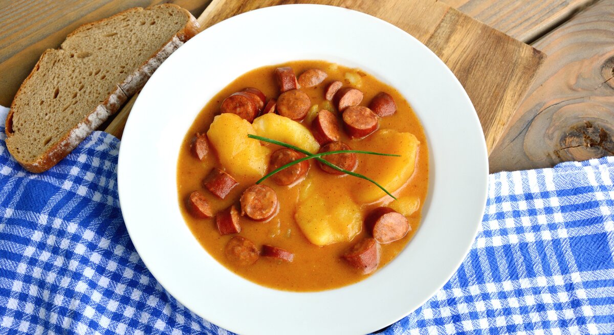 Ein weißer Teller mit der Suppe auf einem Holztisch mit einer blau-weiß-karierten Decke. Alles von schräg oben fotografiert.