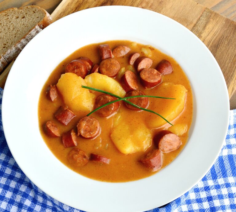 Ein weißer Teller mit der Suppe auf einem Holztisch mit einer blau-weiß-karierten Decke. Alles von schräg oben fotografiert. | © Shutterstock/ PosiNote