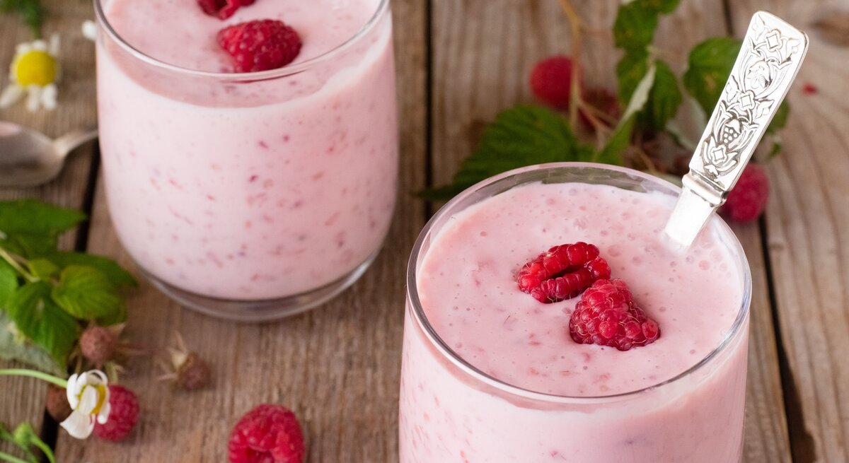 Zwei Gläser mit dem Kokos-Himbeer-Joghurt auf einem rustikalen Holztisch mit frischen Himbeerzweigen.