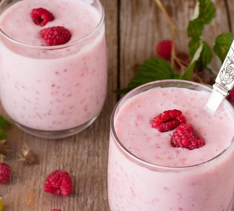 Zwei Gläser mit dem Kokos-Himbeer-Joghurt auf einem rustikalen Holztisch mit frischen Himbeerzweigen. | © Shutterstock/ Yala