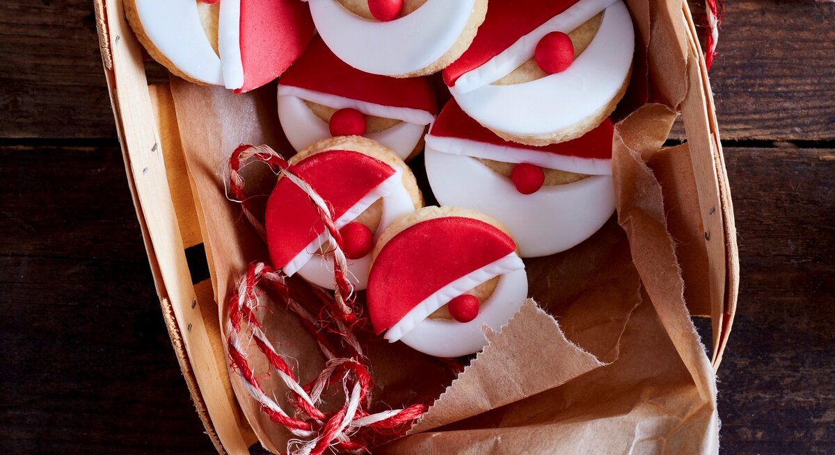 Nikolaus-Kekse auf Krepp in Bastkorb auf dunklem Holztisch aus der Vogelperspektive. Rotes und weißes Fondant mit roter Fondantkugel als Nase ergeben die Gesichter. Rot-weiße Schnur als Deko rechts neben dem Korb und auf dem Korb. 