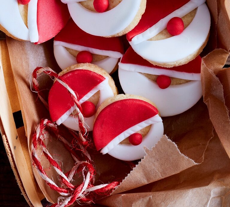 Nikolaus-Kekse auf Krepp in Bastkorb auf dunklem Holztisch aus der Vogelperspektive. Rotes und weißes Fondant mit roter Fondantkugel als Nase ergeben die Gesichter. Rot-weiße Schnur als Deko rechts neben dem Korb und auf dem Korb.  | © Eat Club / Isabell Triemer