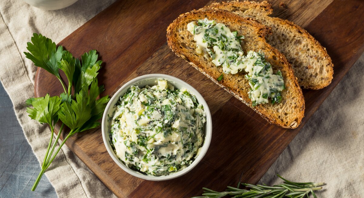Eine Schale vegane Kräuterbutter, daneben zwei Brotscheiben mit der Butter bestrichen, außerdem frische Kräuter. Draufsicht.