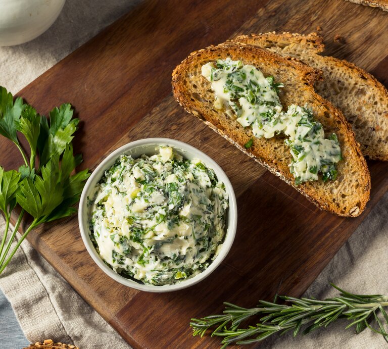 Eine Schale vegane Kräuterbutter, daneben zwei Brotscheiben mit der Butter bestrichen, außerdem frische Kräuter. Draufsicht. | © Shutterstock/ Brent Hofacker