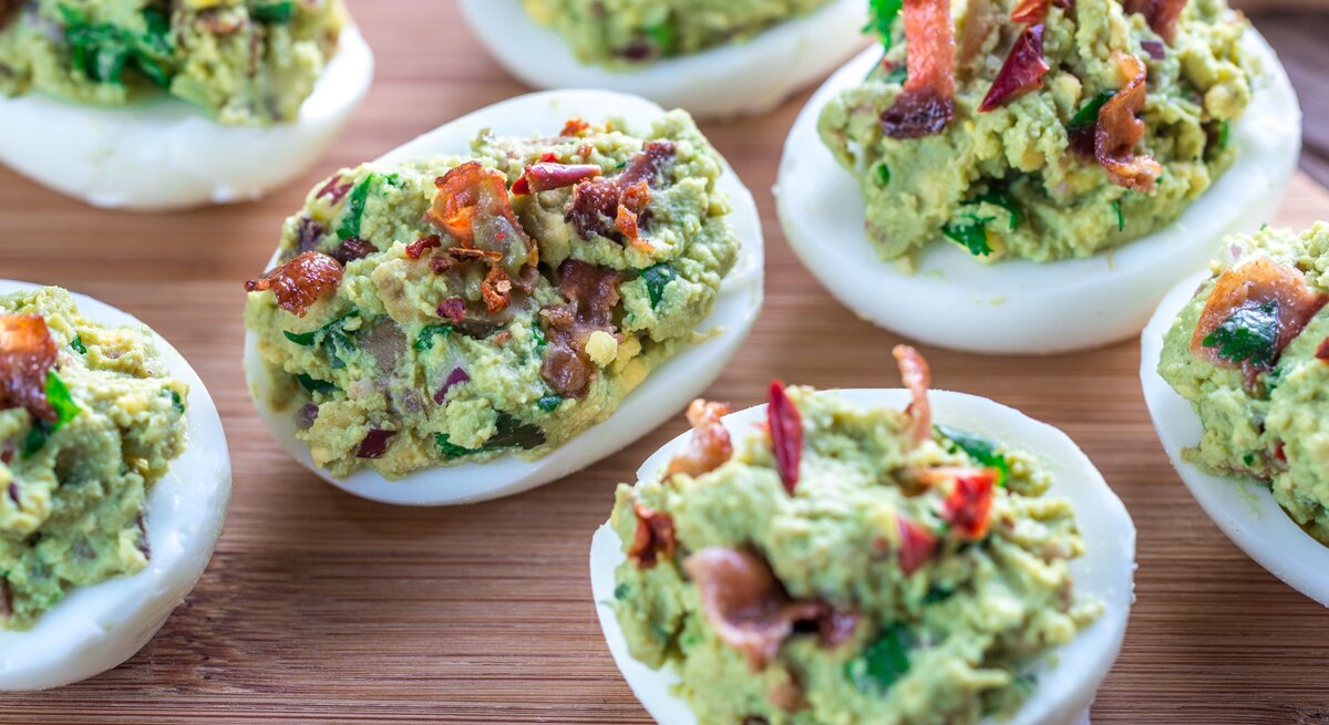 Sieben gefüllte Eier mit Avocado und Bacon auf Holzbrett. Aufnahme aus der Vogelperspektive.