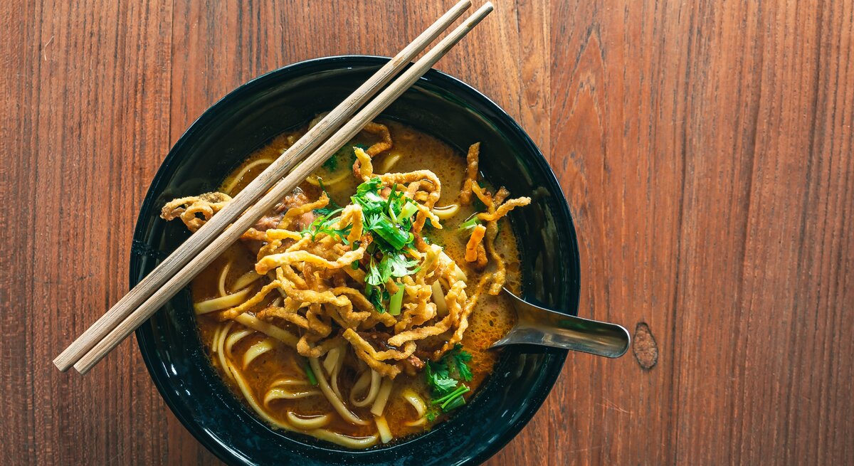 Eine dunkle Schale Khao Soi auf einem dunklen Holztisch mit zwei Holzstäbchen.