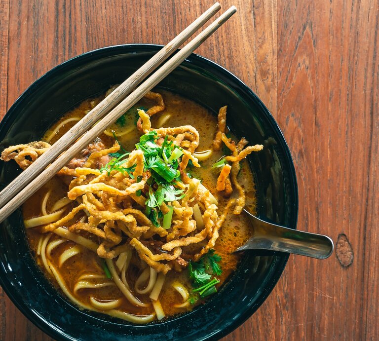 Eine dunkle Schale Khao Soi auf einem dunklen Holztisch mit zwei Holzstäbchen. | © Shutterstock/ YDG