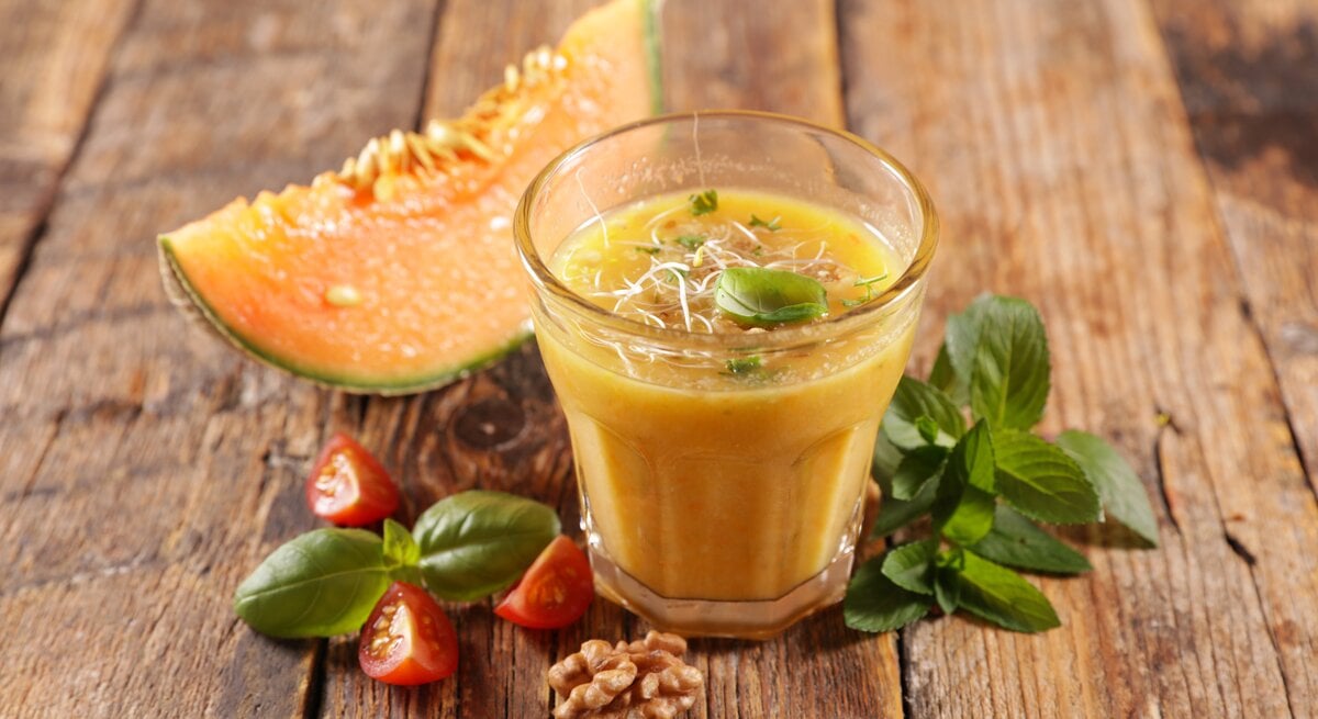 Ein Glas Honigmelonen-Gazpacho mit Sprossen und Basilikum bestreut, daneben liegen ein Stück Melone, Tomaten und Walnüsse.