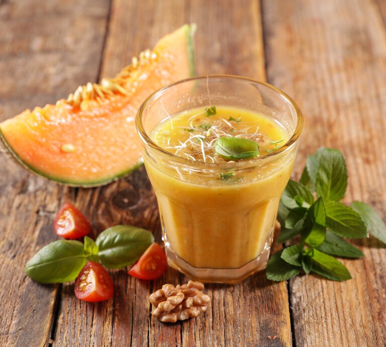 Ein Glas Honigmelonen-Gazpacho mit Sprossen und Basilikum bestreut, daneben liegen ein Stück Melone, Tomaten und Walnüsse. | © Getty Images/ margouillat photo