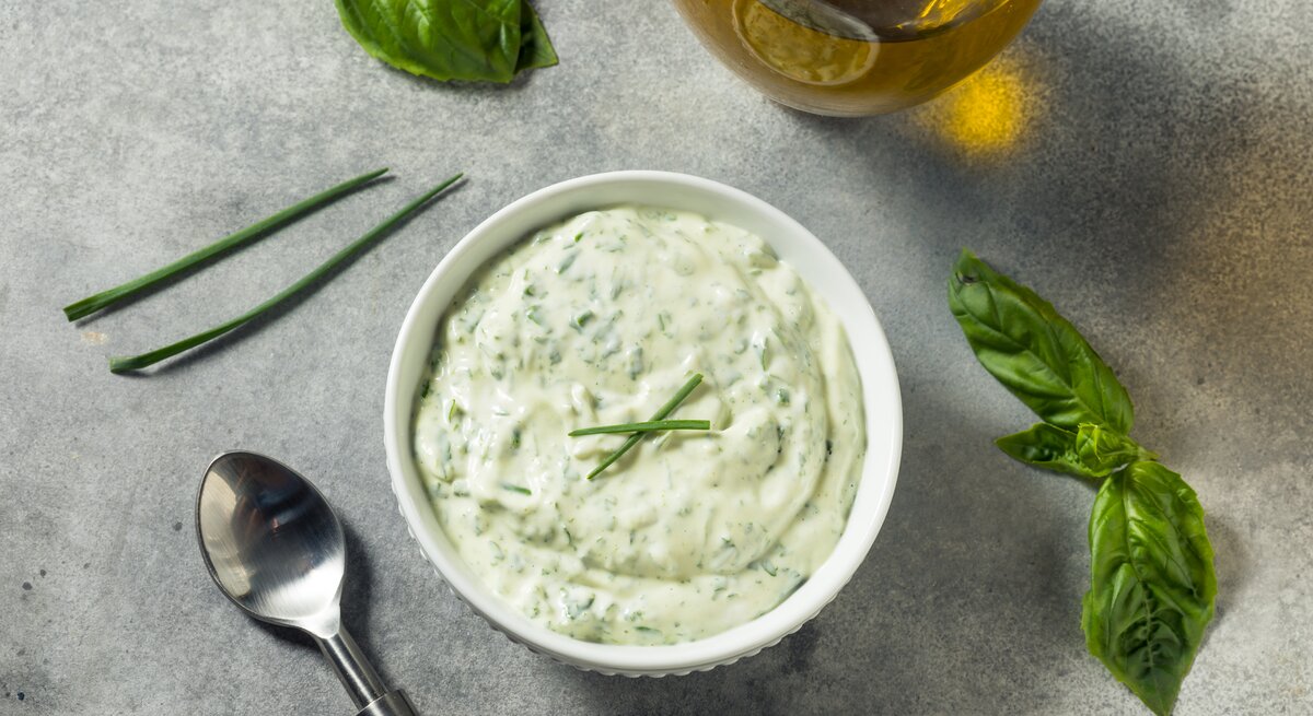 Eine Schale mit Kräuter-Mayonnaise. Daneben steht eine Karaffe Öl und einige Kräuter liegen daneben.