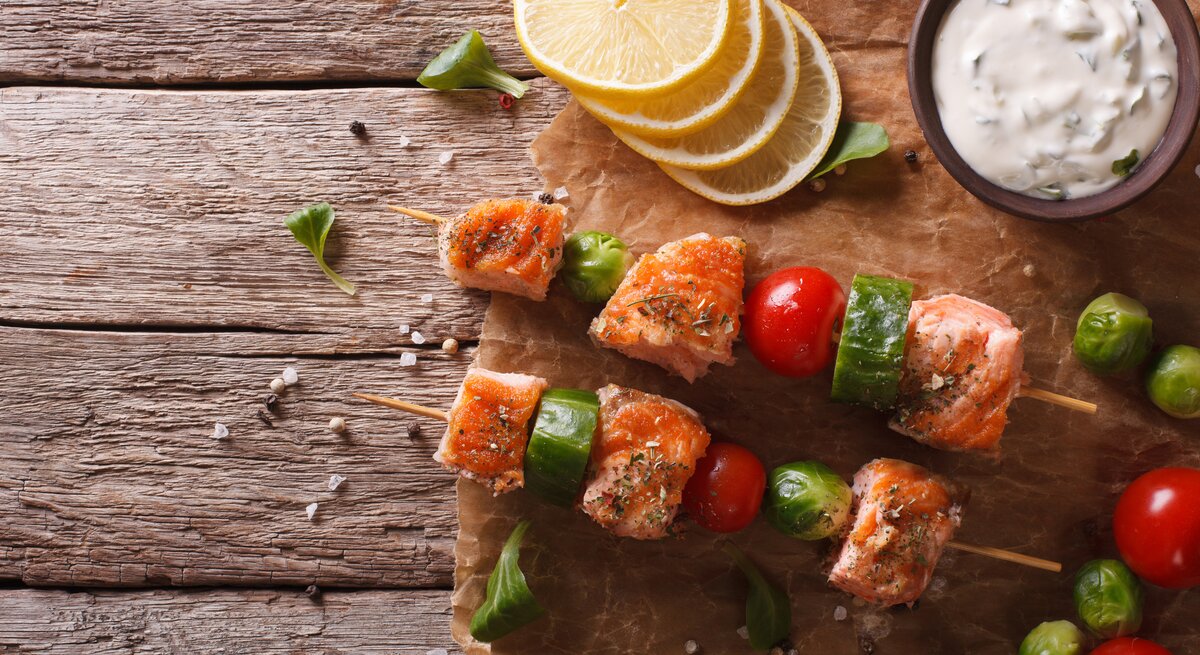 Draufsicht: Zwei Rosenkohl-Lachs-Spieße liegen auf Papier und einem Untergrund aus Holz. Daneben liegen Zitronenscheiben, Rosekohl-Röschen und eine Schale mit Kräuterquark.