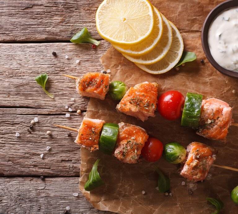 Draufsicht: Zwei Rosenkohl-Lachs-Spieße liegen auf Papier und einem Untergrund aus Holz. Daneben liegen Zitronenscheiben, Rosekohl-Röschen und eine Schale mit Kräuterquark. | © Shutterstock / Sergii Koval