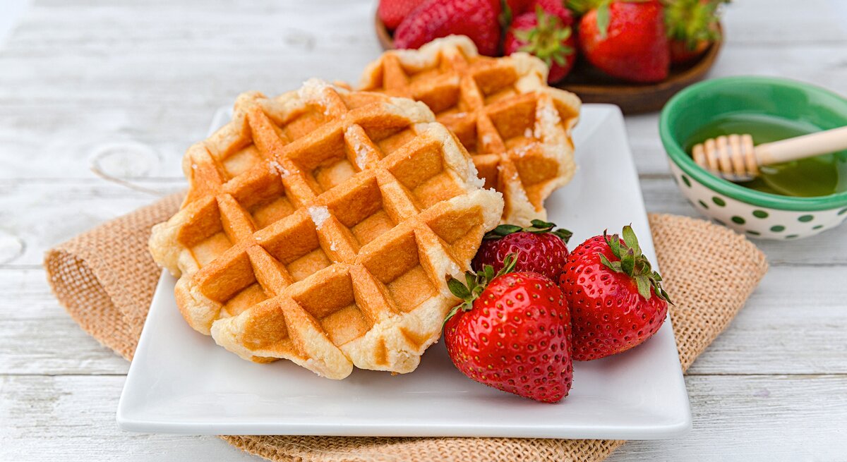 Frontal: Weißer Untergund aus Holt. Darauf liegt ein grobes leinentcuh mit einem weizen Teller. Auf dem sind zwei Haferkleie-Waffeln mit frischen Erdbeeren. Im Hintergrund sind weitere frische Erdbeeren, sowie ein Schälchen mit Honig.