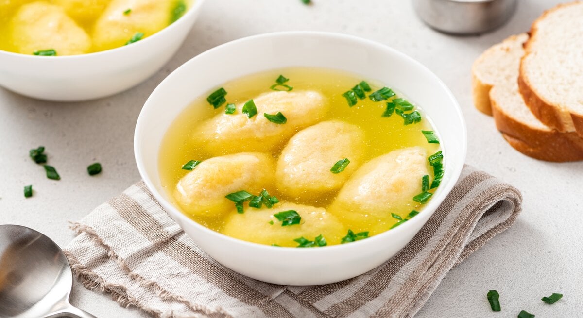 Draufsicht, schräg: Zwei weiße Schüsseln mit einer bayerischen Grießnockerlsuppe stehen auf einem hellen Untergrund. Daneben liegen ein Löffel, zwei Scheiben Weißbrot und eine Schale mit Salz.