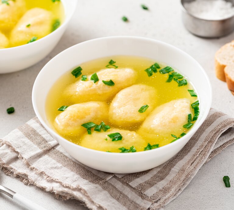 Draufsicht, schräg: Zwei weiße Schüsseln mit einer bayerischen Grießnockerlsuppe stehen auf einem hellen Untergrund. Daneben liegen ein Löffel, zwei Scheiben Weißbrot und eine Schale mit Salz. | © Shutterstock / Svetlana Monyakova