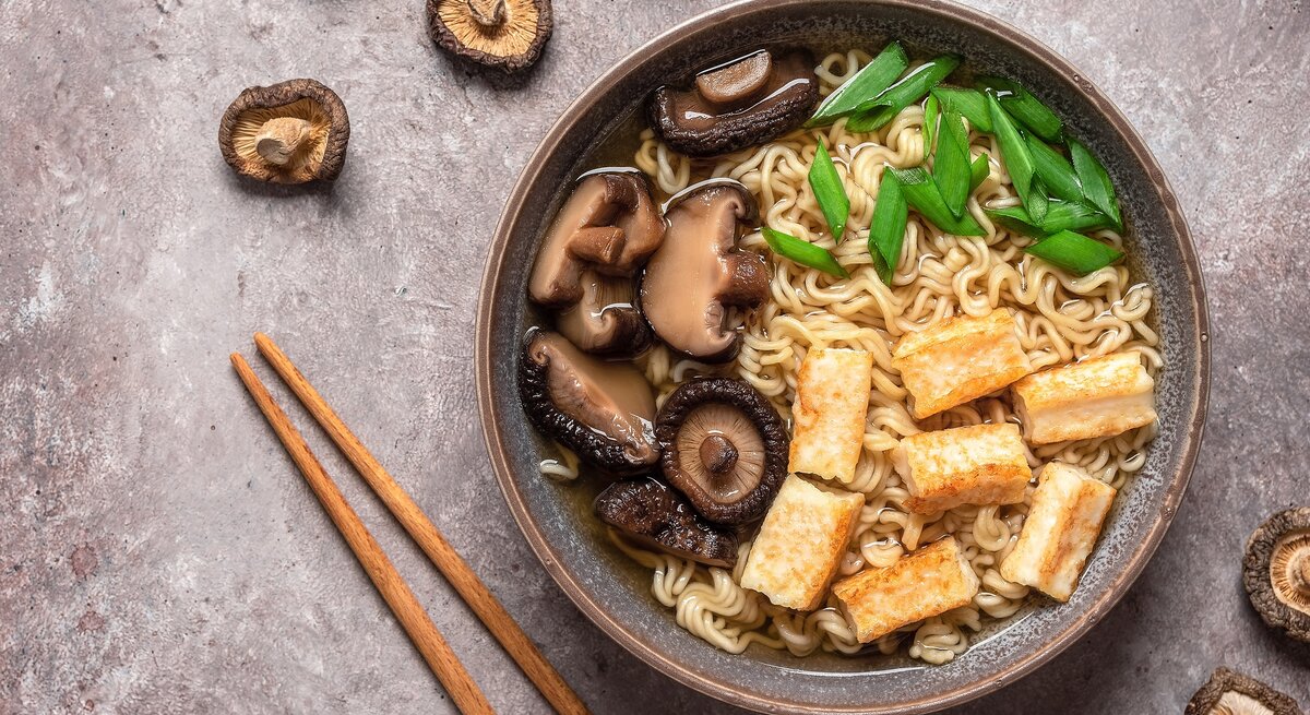 Eine Schüssel schnelle Ramen mit Nudeln, Tofu, Pilzen und Frühlingszwiebeln in der Draufsicht, darum herum liegen Pilze und Essstäbchen.