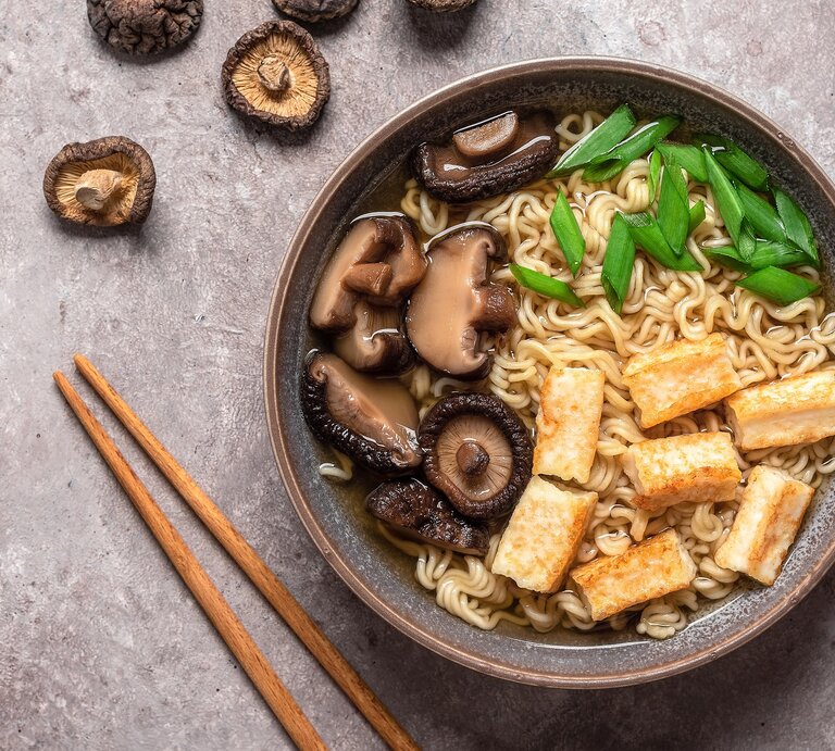 Eine Schüssel schnelle Ramen mit Nudeln, Tofu, Pilzen und Frühlingszwiebeln in der Draufsicht, darum herum liegen Pilze und Essstäbchen. | © Adobe Stock/ Yulia