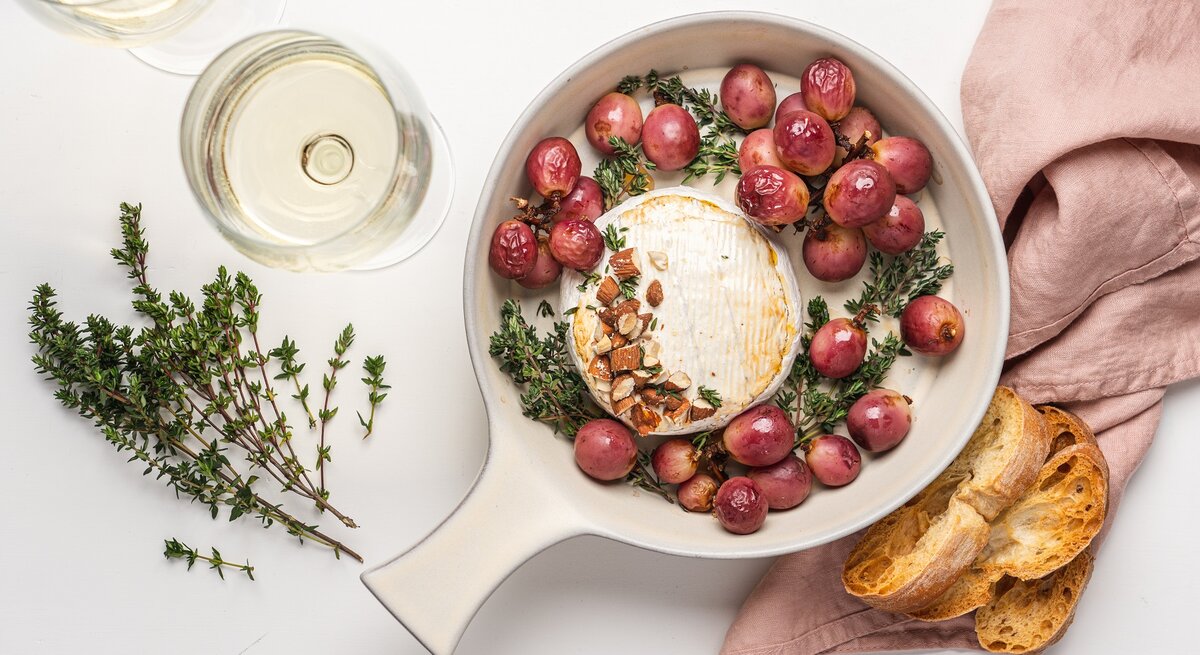 Eine feuerfeste Form mit einem Ofenkäse mit Weintrauben, Thymian, Honig und Nüssen, daneben stehen zwei Gläser mit Weißwein und einer altrosa Stoff-Serviette zur Dekoration.