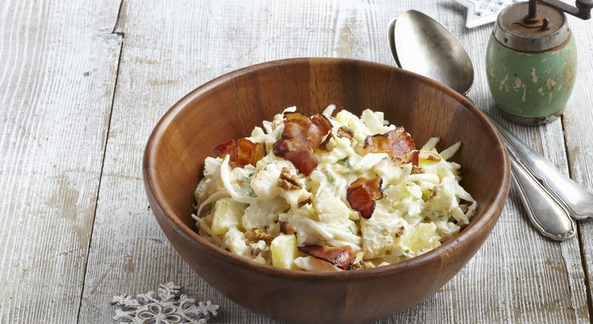 Auf einem Untergund aus Holz steht eine Schüssel aus Holz. In der ist ein Kartoffelsalat mit Blumenkohl, Walnüssen und Speck. Daneben stehen eine Pfeffermühle, zwei Löffel sowie zwei Sterne.