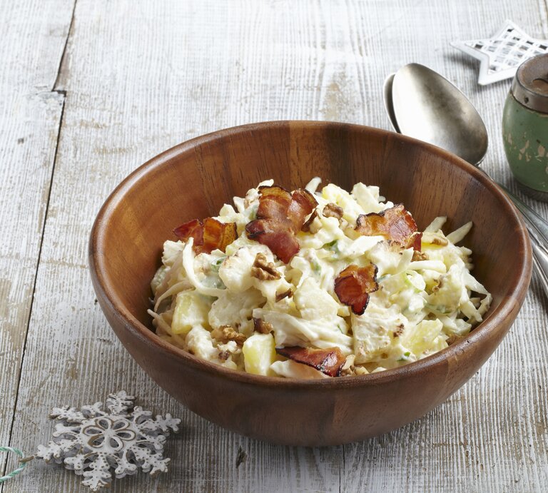 Auf einem Untergund aus Holz steht eine Schüssel aus Holz. In der ist ein Kartoffelsalat mit Blumenkohl, Walnüssen und Speck. Daneben stehen eine Pfeffermühle, zwei Löffel sowie zwei Sterne. | © StockFood / Profimedia